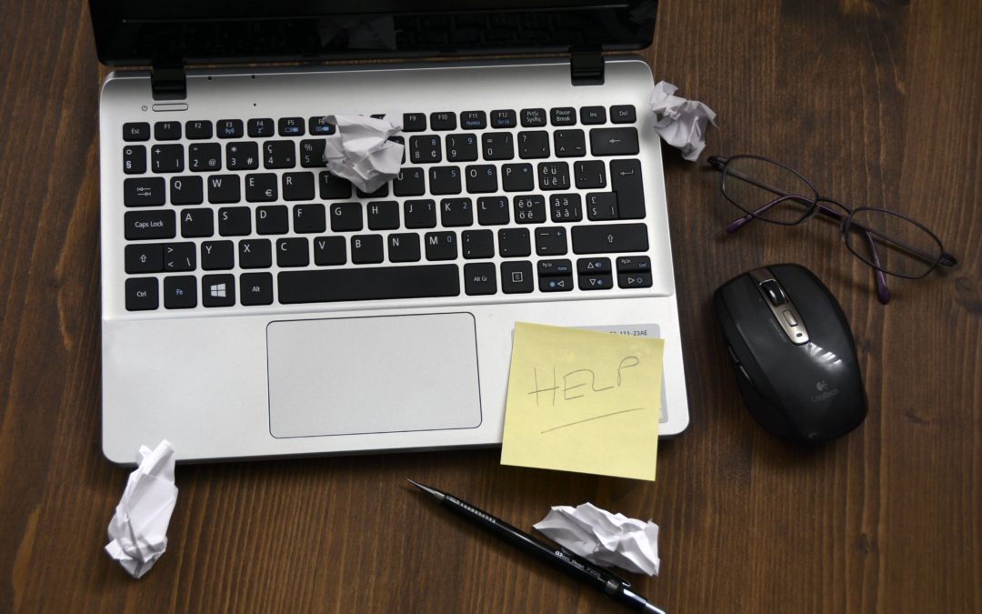 A silver Windows laptop computer sits on a dark brown wooden desk. Several balled up scraps of paper and a sticky note with "HELP" written on it are scattered around the frame.