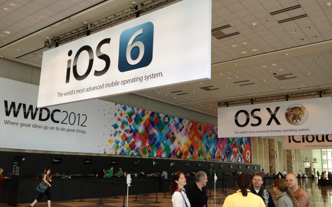 An interior view of a convention hall with promotional banners. One reads "iOS 6" and another reads "OS X." The wall reads "WWDC 2012."