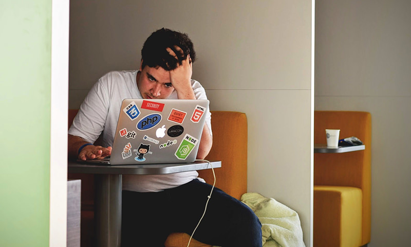 computer virus removal in Cordova, Alaska by CDV Tech Guy A young man in a white t-shirt and jeans sits in a restaurant booth. He is looking at his laptop in frustration with his left hand in his hair. The laptop has been personalized with an array of stickers.